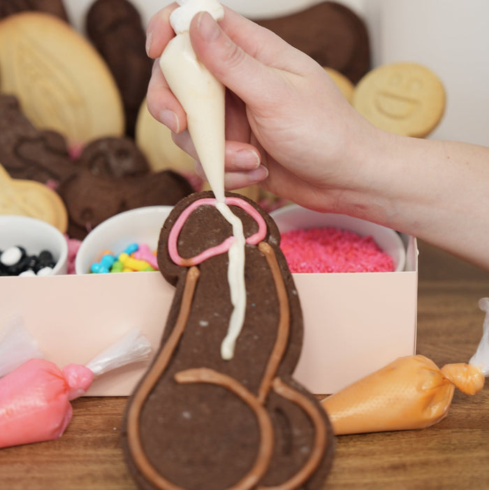 Person decorating a chocolate cookie with white icing on a wooden table.