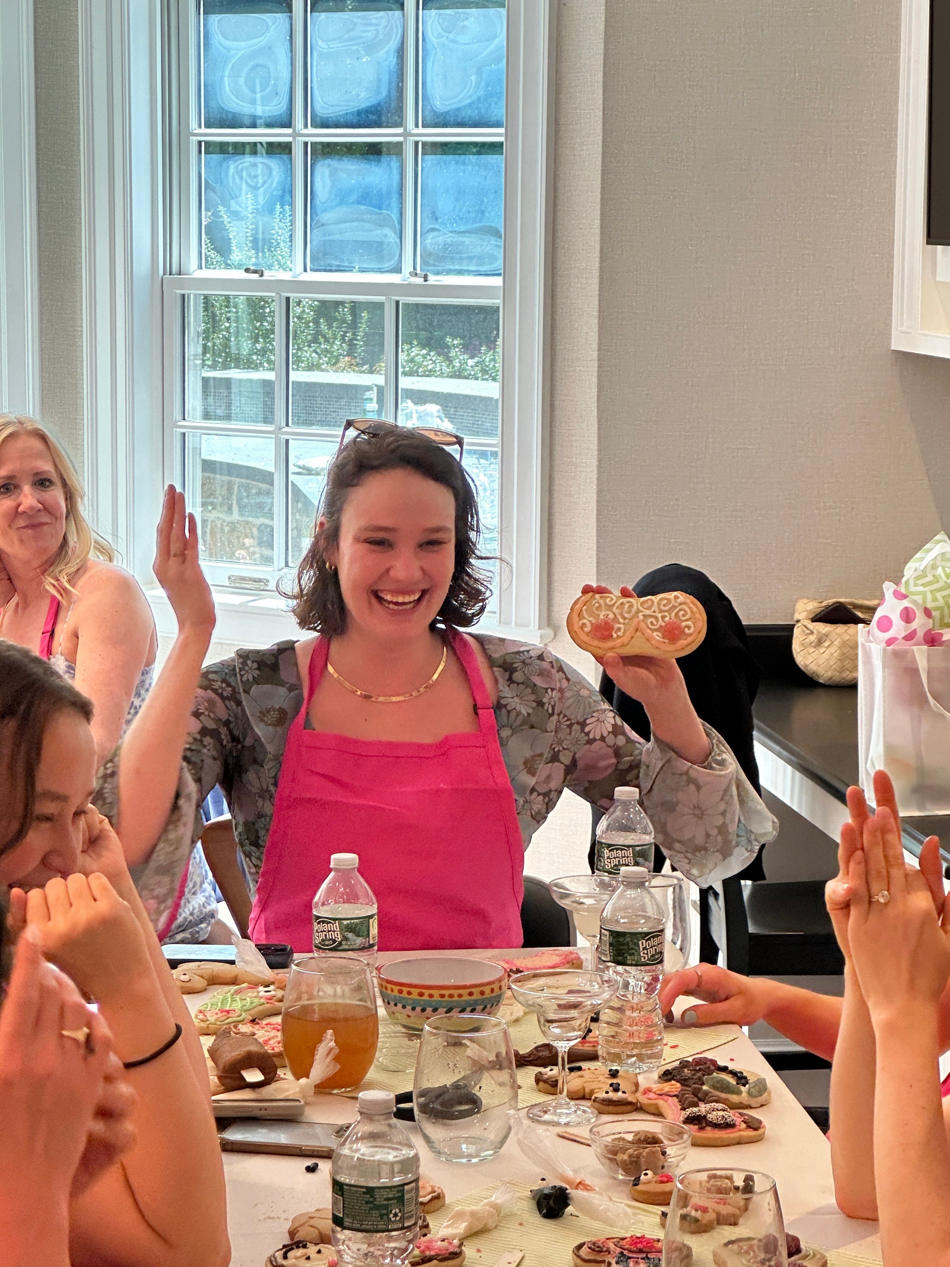 white woman in a pink apron smiling at the camera holding up breast-shaped cookies at a party